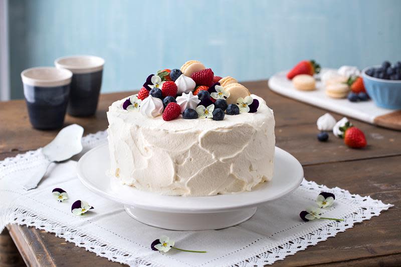 Klassisk bløtkake med hjemmelaget vaniljekrem pyntet med bringebær, blåbær, pikekyss, makroner og blomster. Kaken står på rustikk trebord, med hvit blondeduk.