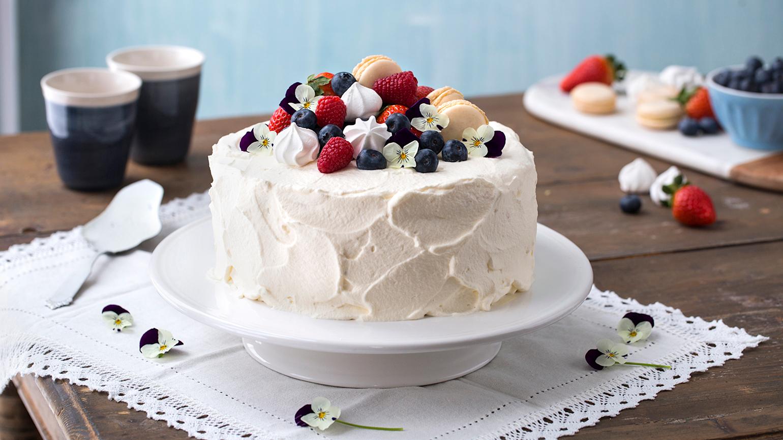 Klassisk bløtkake med hjemmelaget vaniljekrem pyntet med bringebær, blåbær, pikekyss, makroner og blomster. Kaken står på rustikk trebord, med hvit blondeduk.