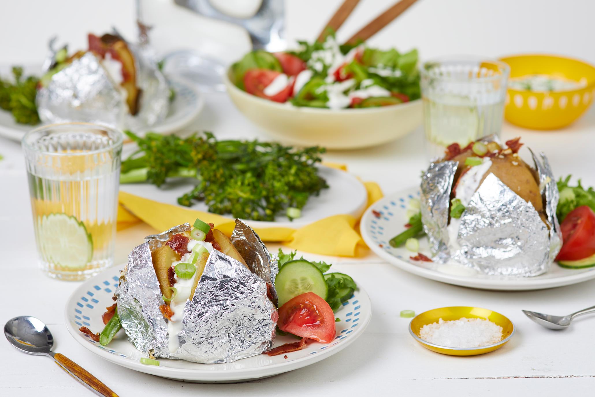 Bakepoteter med bacon og urtedressing på to hvite tallerkener foran et fat med brokkolinei og en bolle med salat