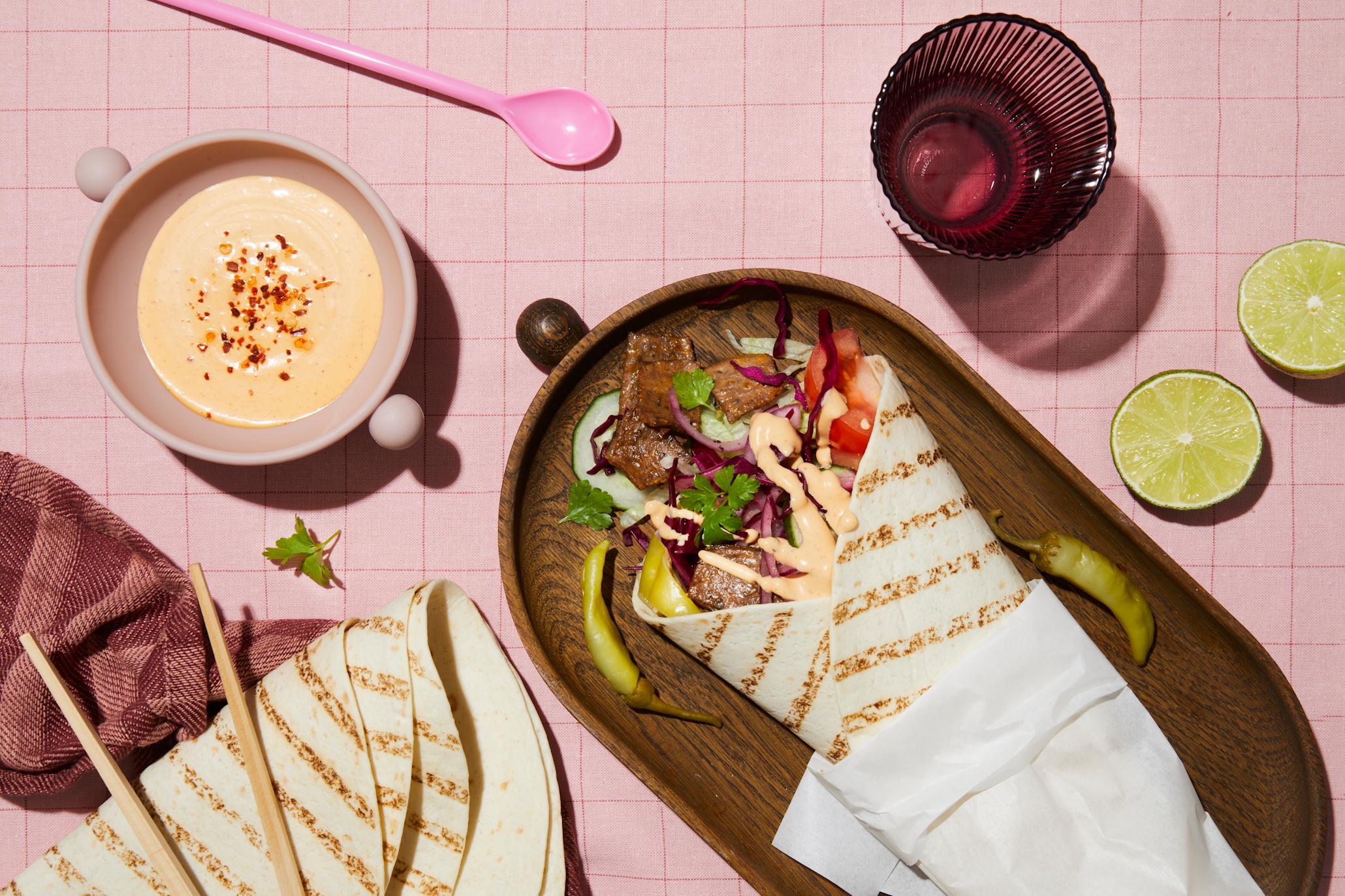 Rullekebab med syltet rødløk og rødkål, syltet chili, grønnsaker og kebabsaus, servert i wrap på mørkt trefat på rosa rutet duk. Halve lime, og liten skål med kebabsaus på siden.
