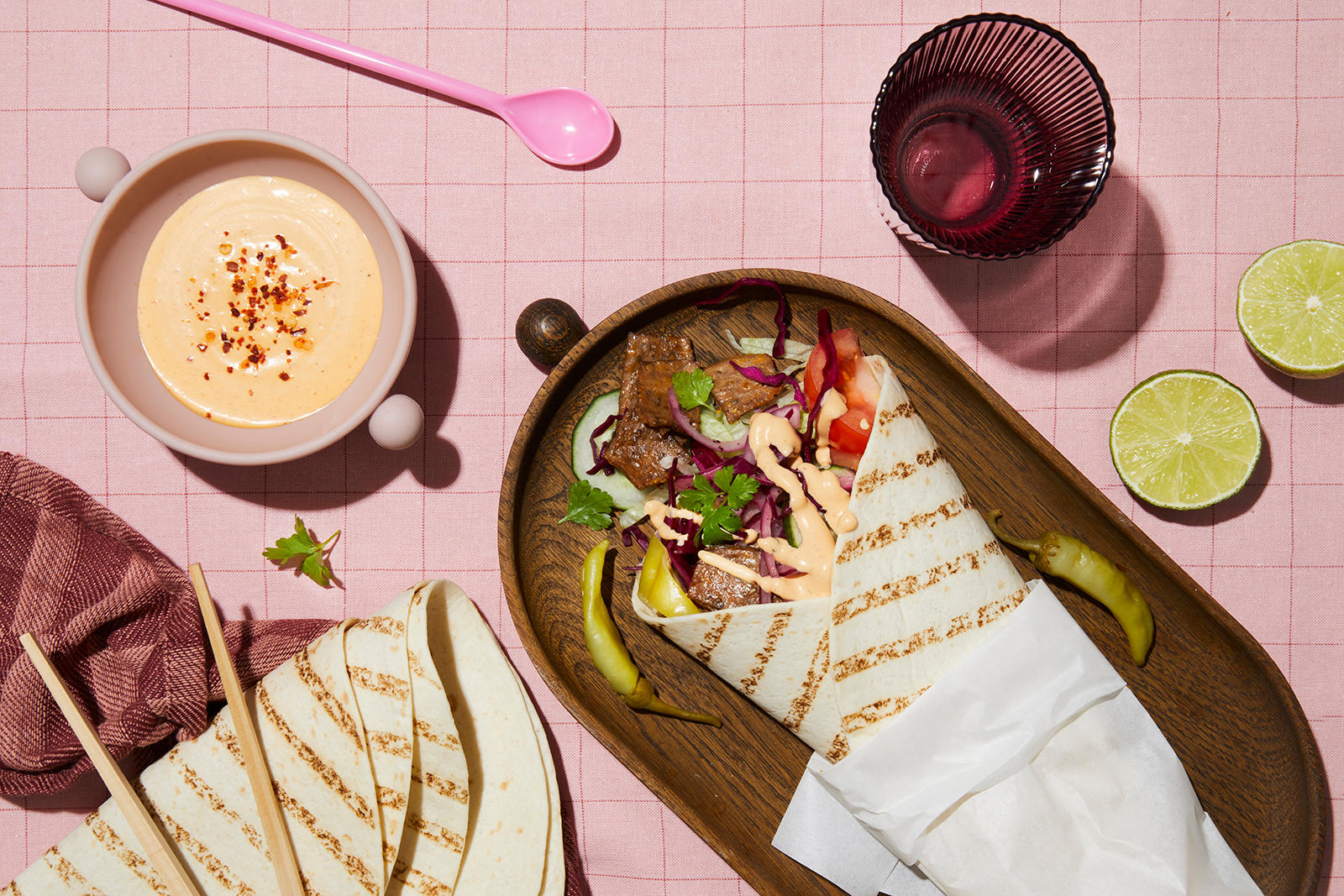 Rullekebab med syltet rødløk og rødkål, syltet chili, grønnsaker og kebabsaus, servert i wrap på mørkt trefat på rosa rutet duk. Halve lime, og liten skål med kebabsaus på siden.