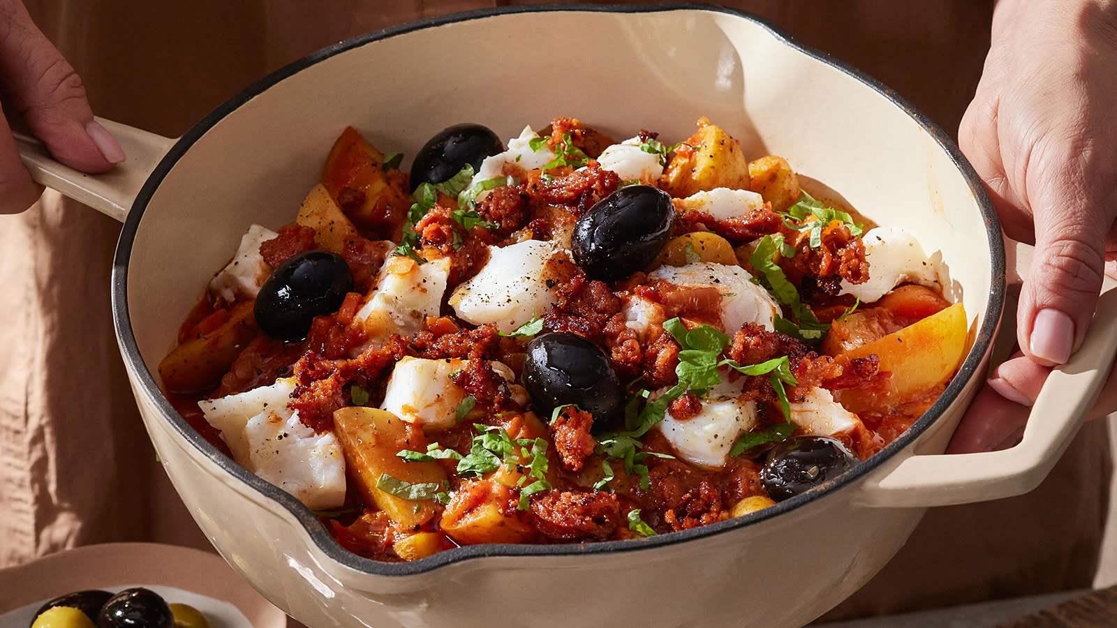 Bacalao med torsk, chorizo og poteter servert i hvit emaljert støpejernsgryte, med svarte oliven og friske urter på toppen.