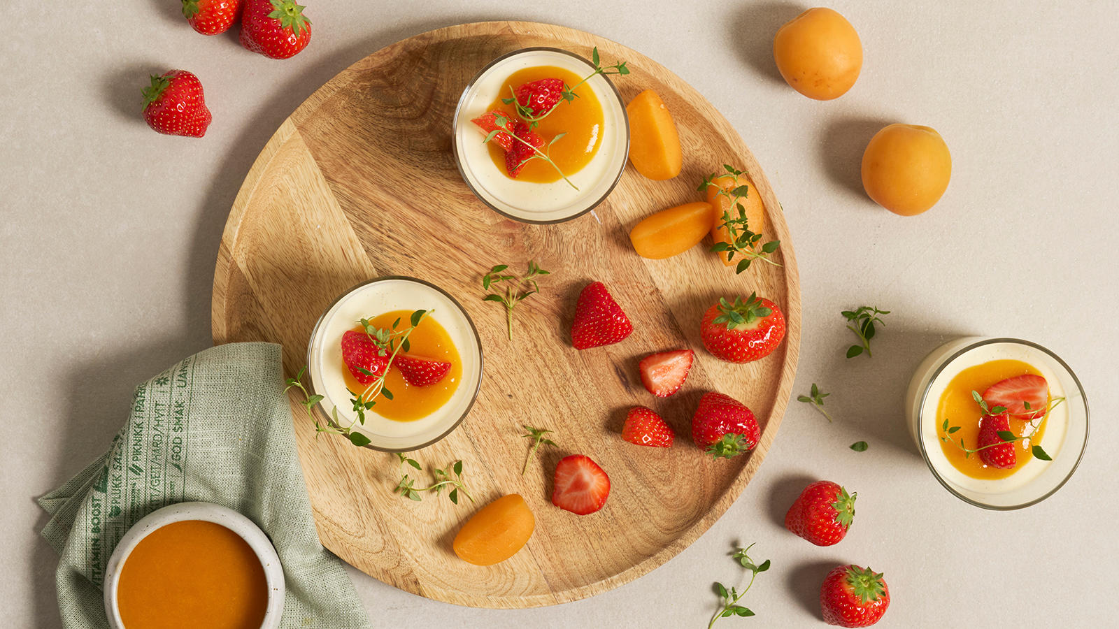 Panna cotta med aprikostopping servert i små dessertglass med friske jordbær og kvister med timian. Glassene står på lyst rundt trefat.