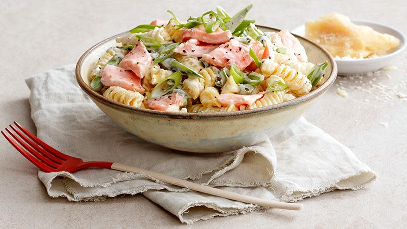 Kremet pastasalat i lys bolle, med laks, sukkererter, og vårløk. Lys bakgrunn med beige linserviett og rød gaffel. Parmesan i bakgrunnen.
