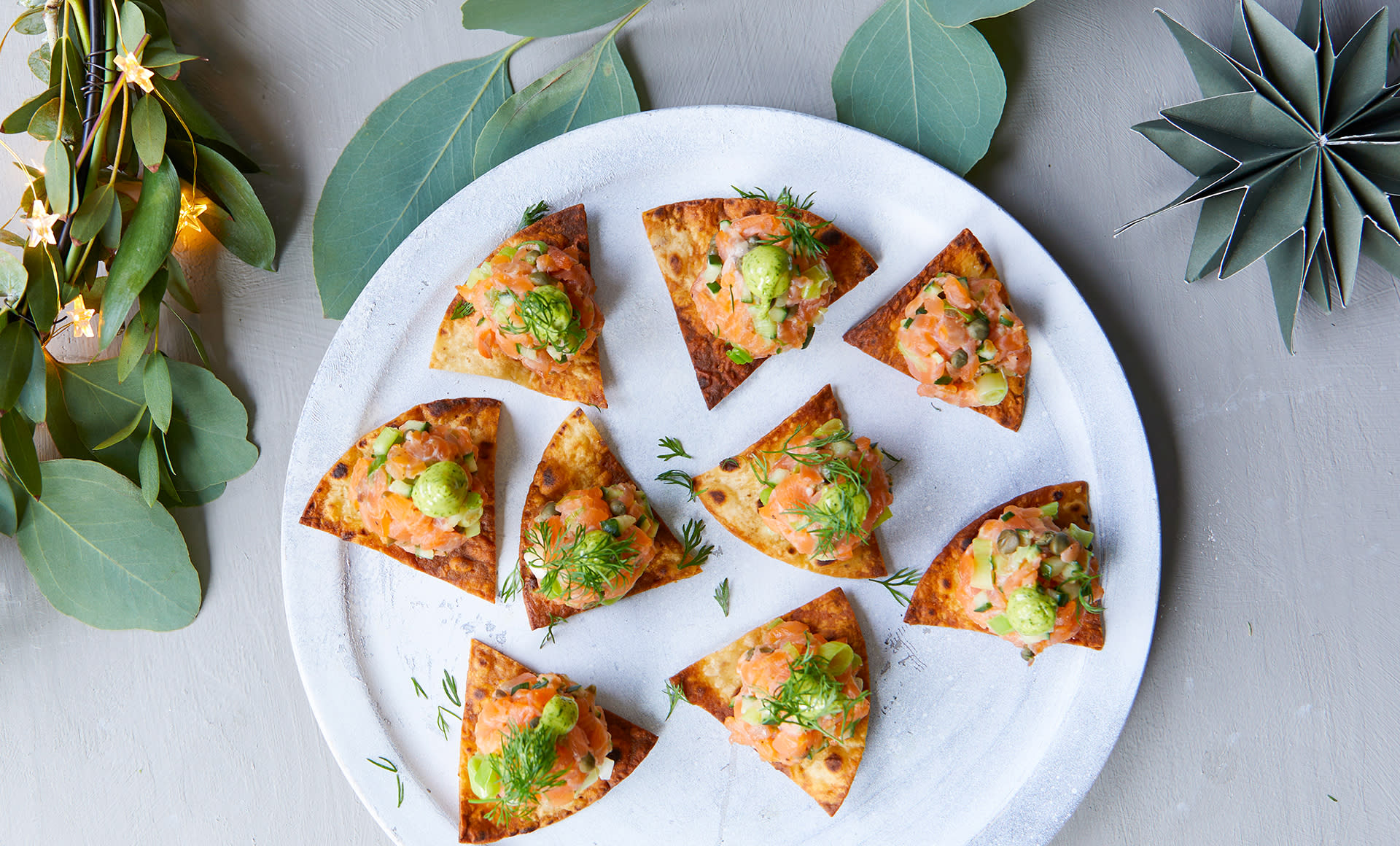 Røkelakstartar på lompechip servert på hvit tallerken med dillmajones og frisk dill på toppen. Grønn julepynt og julelys rundt tallerkenen.