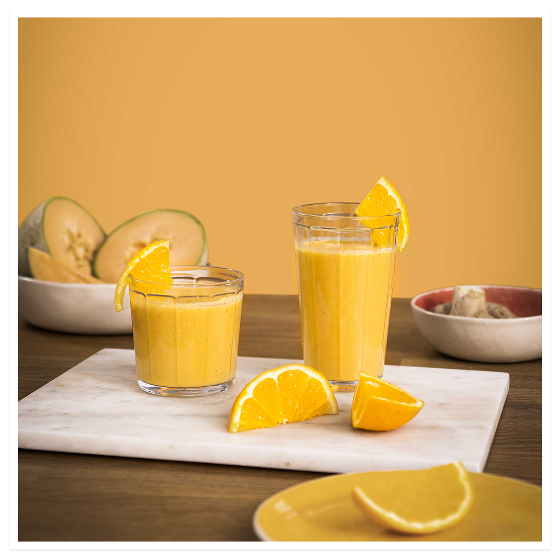 To stykk Glød -smoothier i glass står på hvit marmorfjøl på mørkt trebord, med cantaloupe melon i bakgrunnen, ferske appelsinbåter og ingefær.