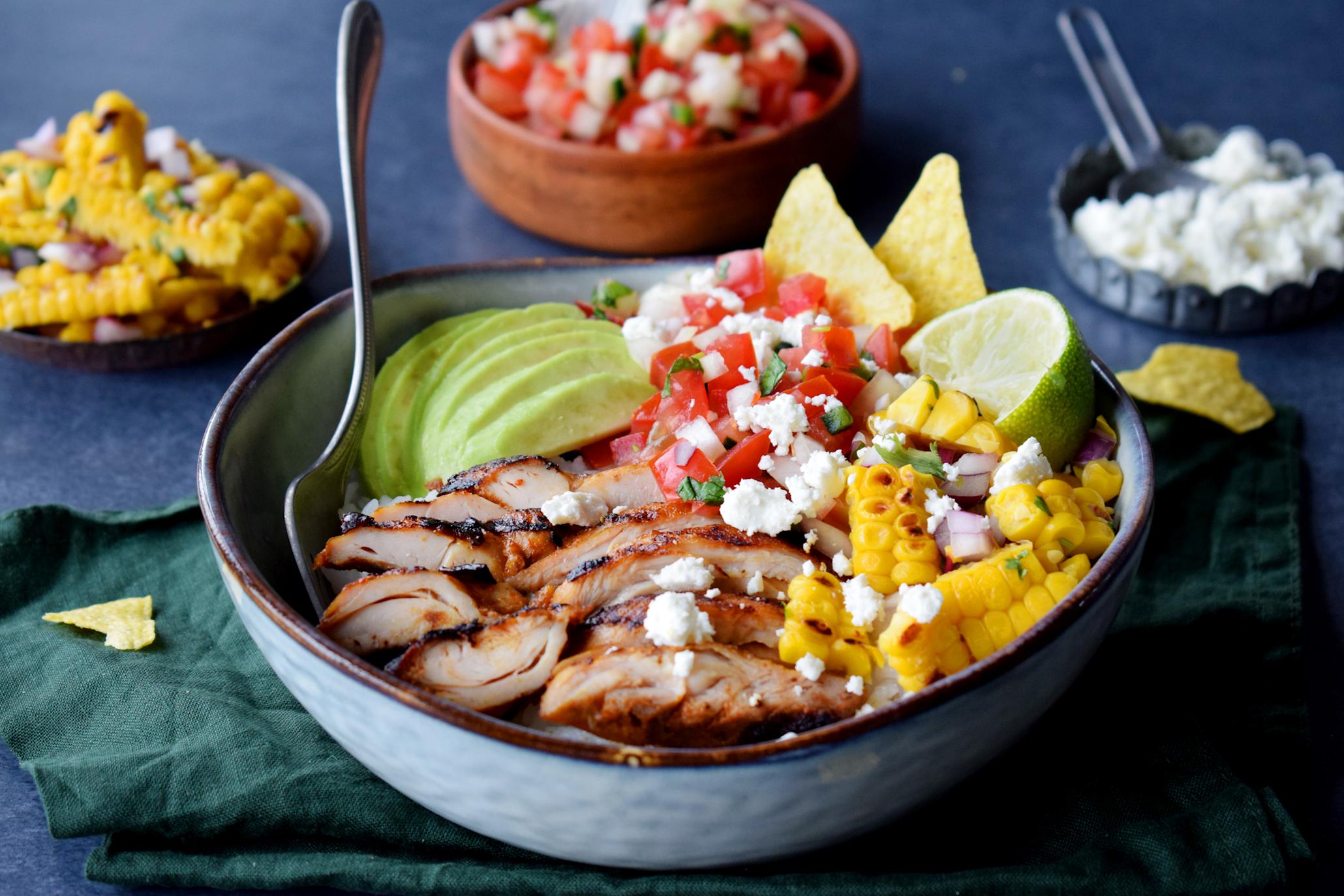 Burrito bowl med kylling, pico de gallo og grillet maissalsa, servert med tortilla chips og feta ost i blå keramikkskål på mørk bakgrunn. Skåler med maissalsa, pico de gallo og fetaost i bakgrunnen.