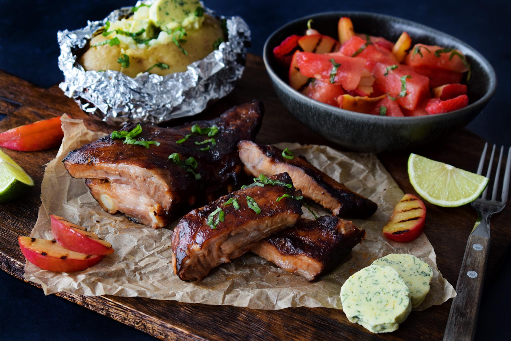Baby back ribs med bakt potet, hjemmelaget hvitløkssmør, og vannmelonsalat med fersken servert på mørk trefjøl.