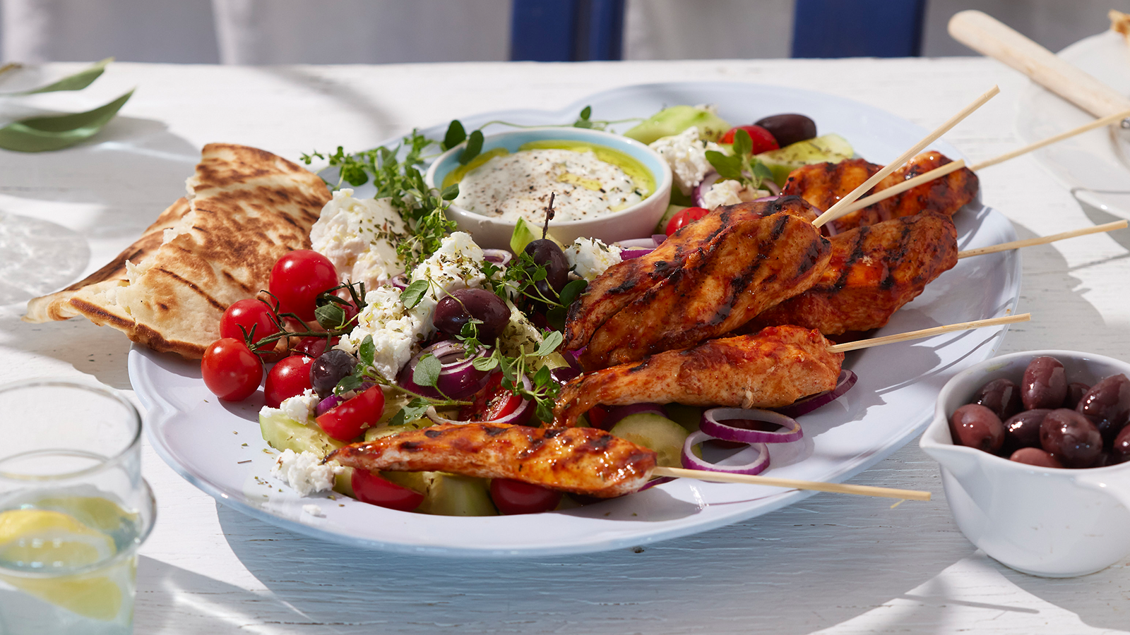 Grillspyd med pita og gresk salat servert på lys avlang asjett med tzatziki, agurk, tomater, fetaost og oliven.