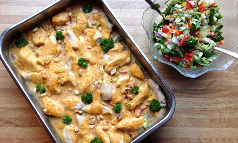 Biter av kyllingfilet i form sammen med rødløk og purre, red curry og kokosmelk, på trebord. Frisk salat ved siden av.