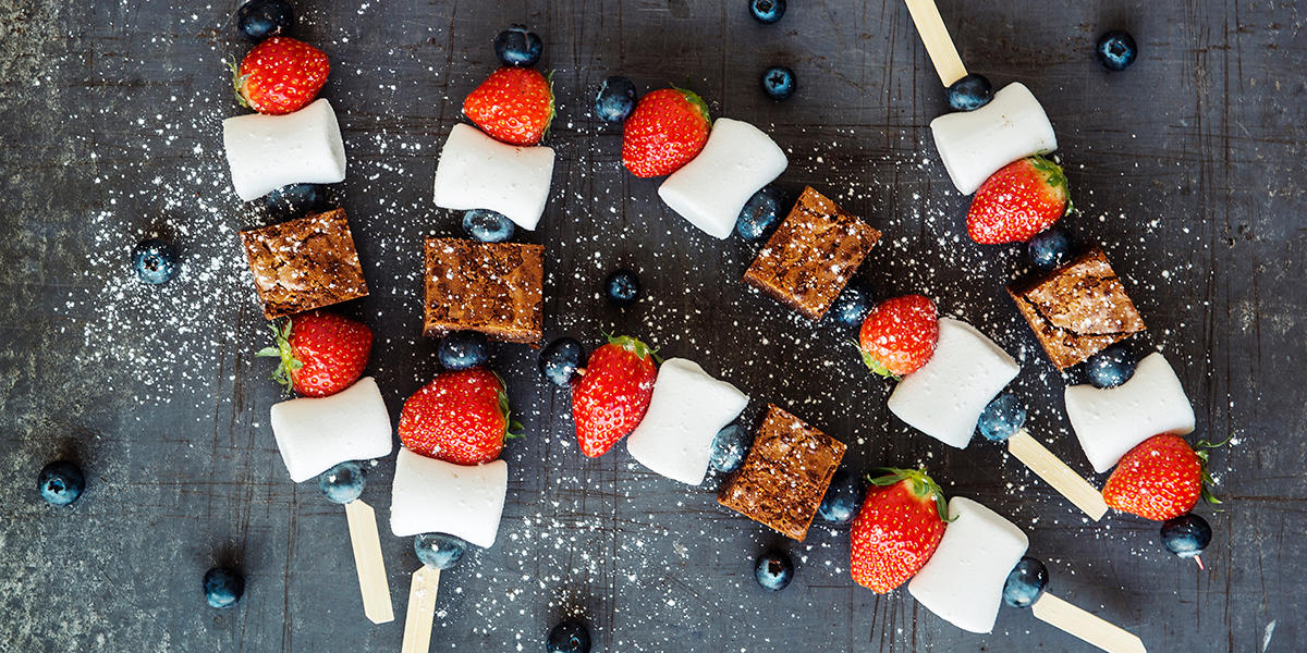 Browniespyd med ferske jordbær, blåbær og marshmallows servert med et dryss melis på mørk bakgrunn. Fargene i bærene og marshmallowsen representerer fargene i flagget i anledning 17. Mai.