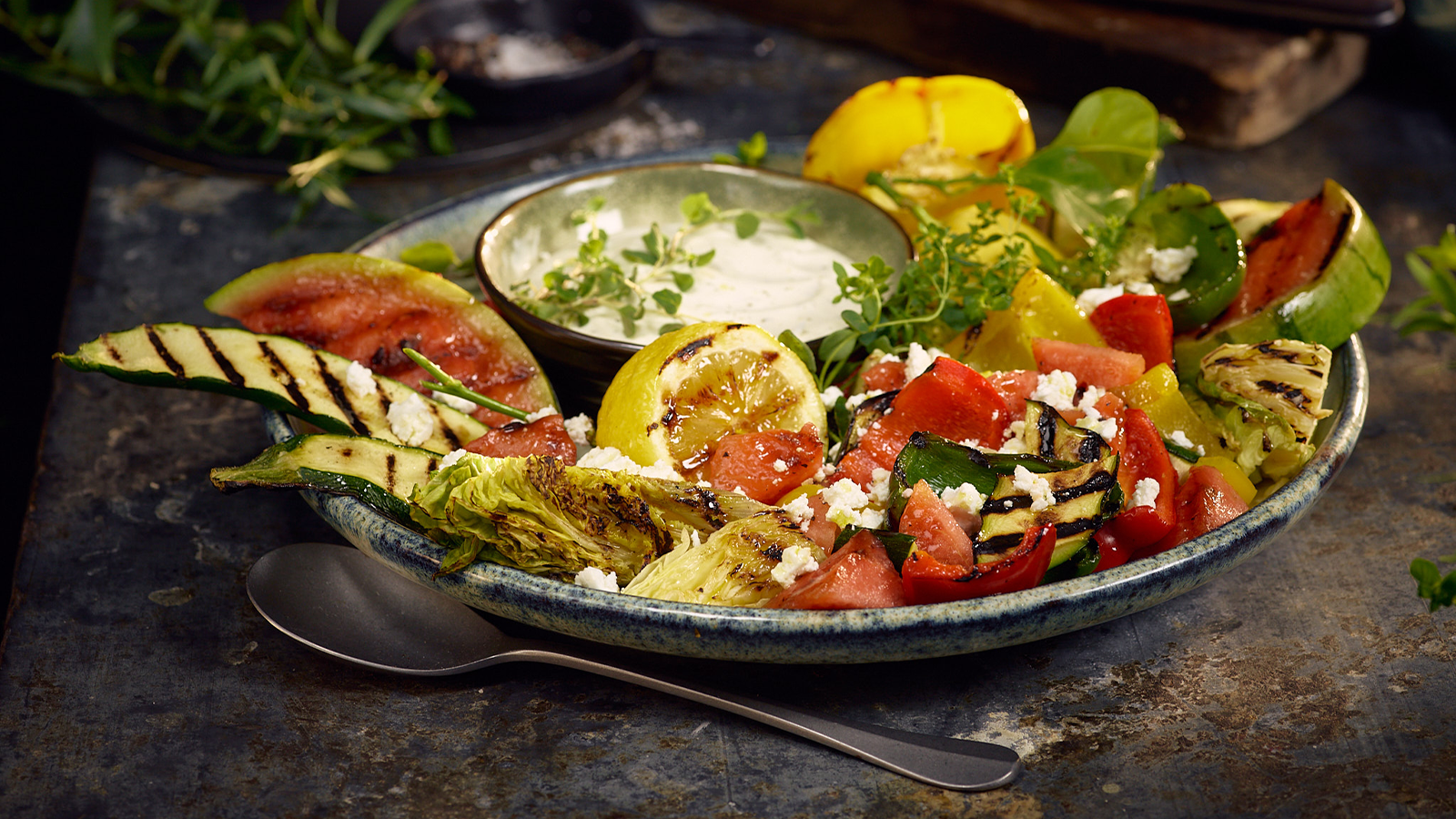 Grillet sommersalat med feta og hvitløksdressing servert på keramikktallerken med skje ved siden av.