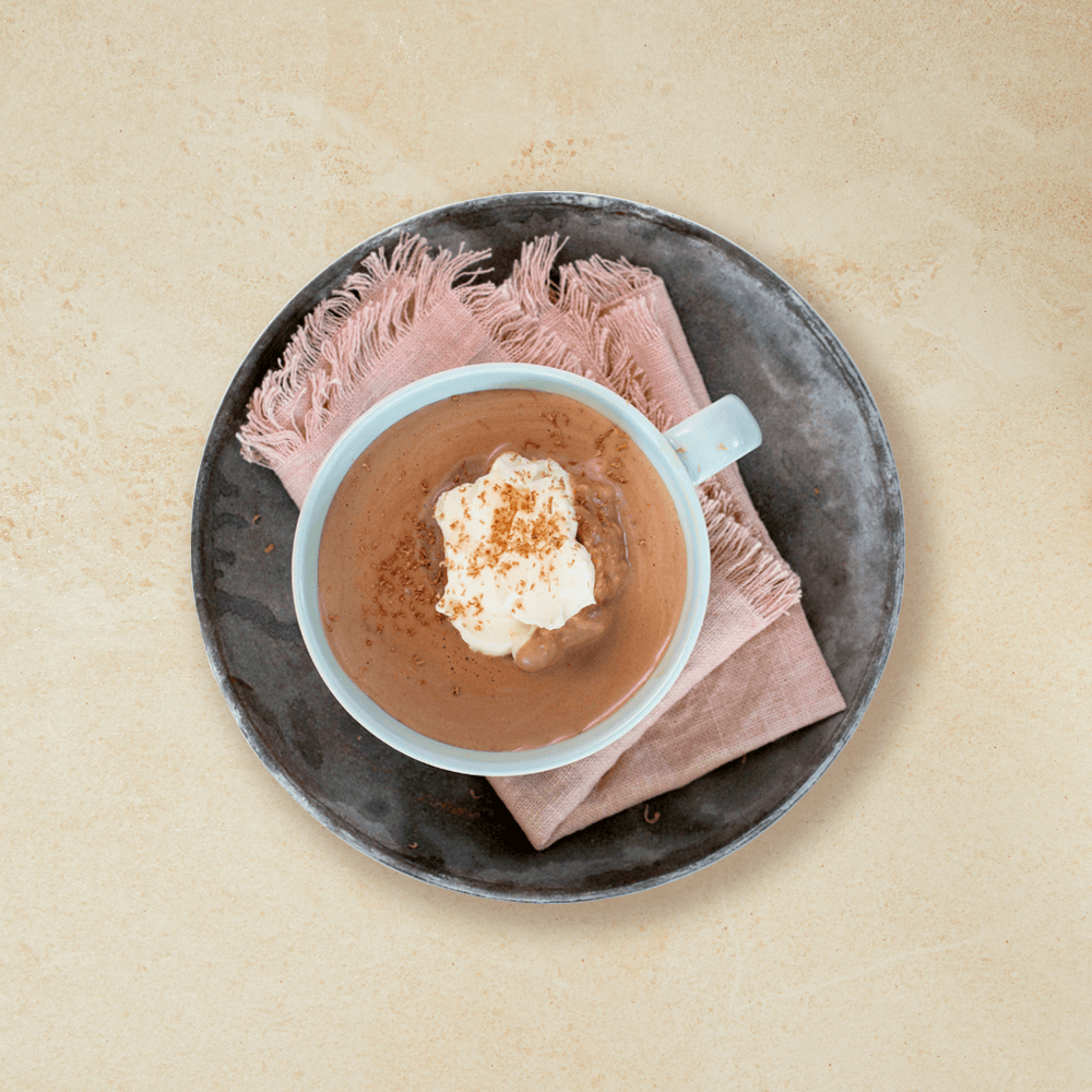 Varm melkesjokolade med krem i en lys blå kopp, servert på lyserosa stoffserviett på mørkegrå asjett. Beige bakgrunn.