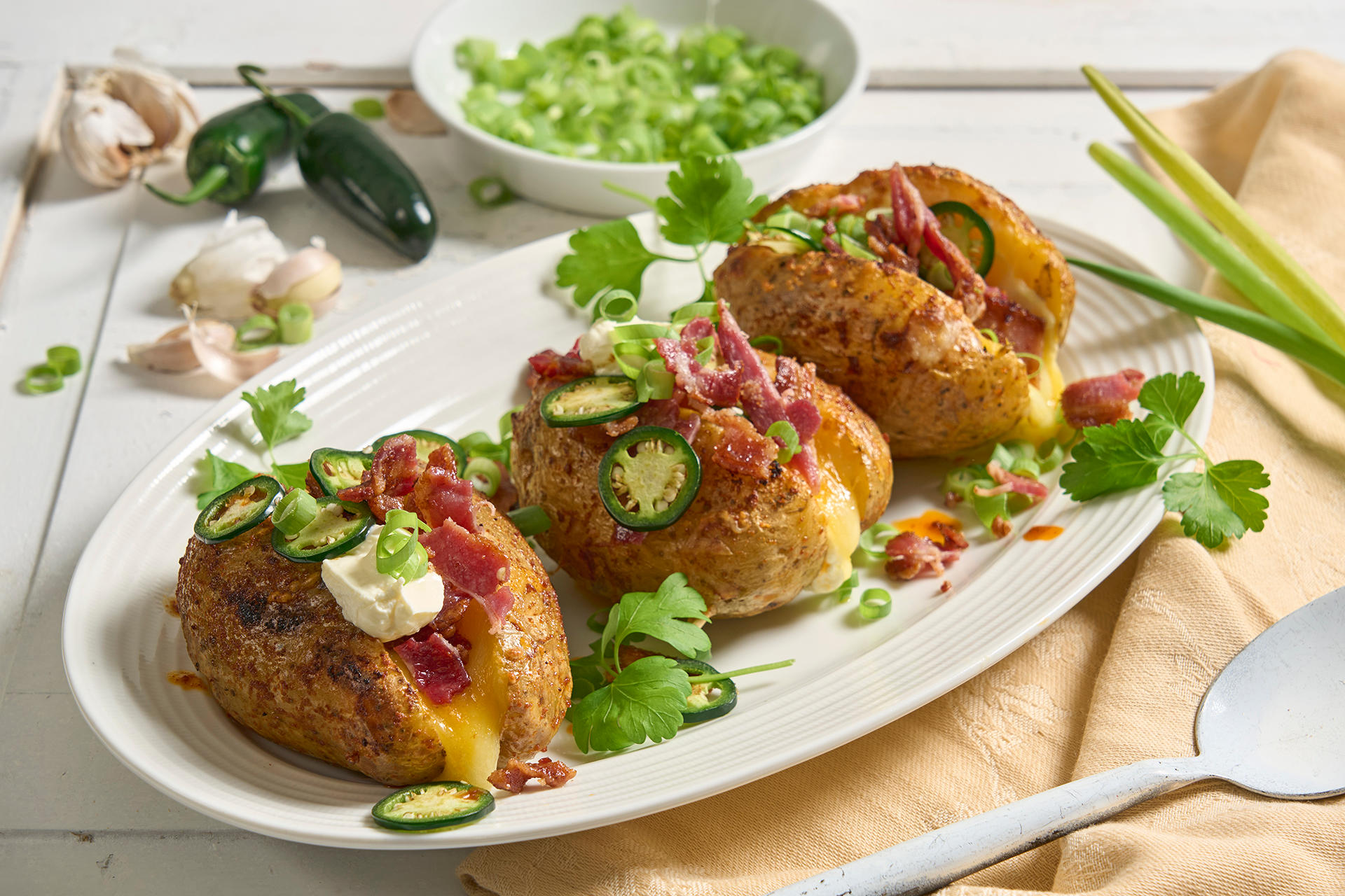 Cheesy bakt potet på grillen med bacon og jalapeño, toppet med grønne urter, kremost og smeltet ost, servert på ovalt hvitt fat. Jalapeño, hvitløk og en skål finsnittet vårløk i bakgrunnen.