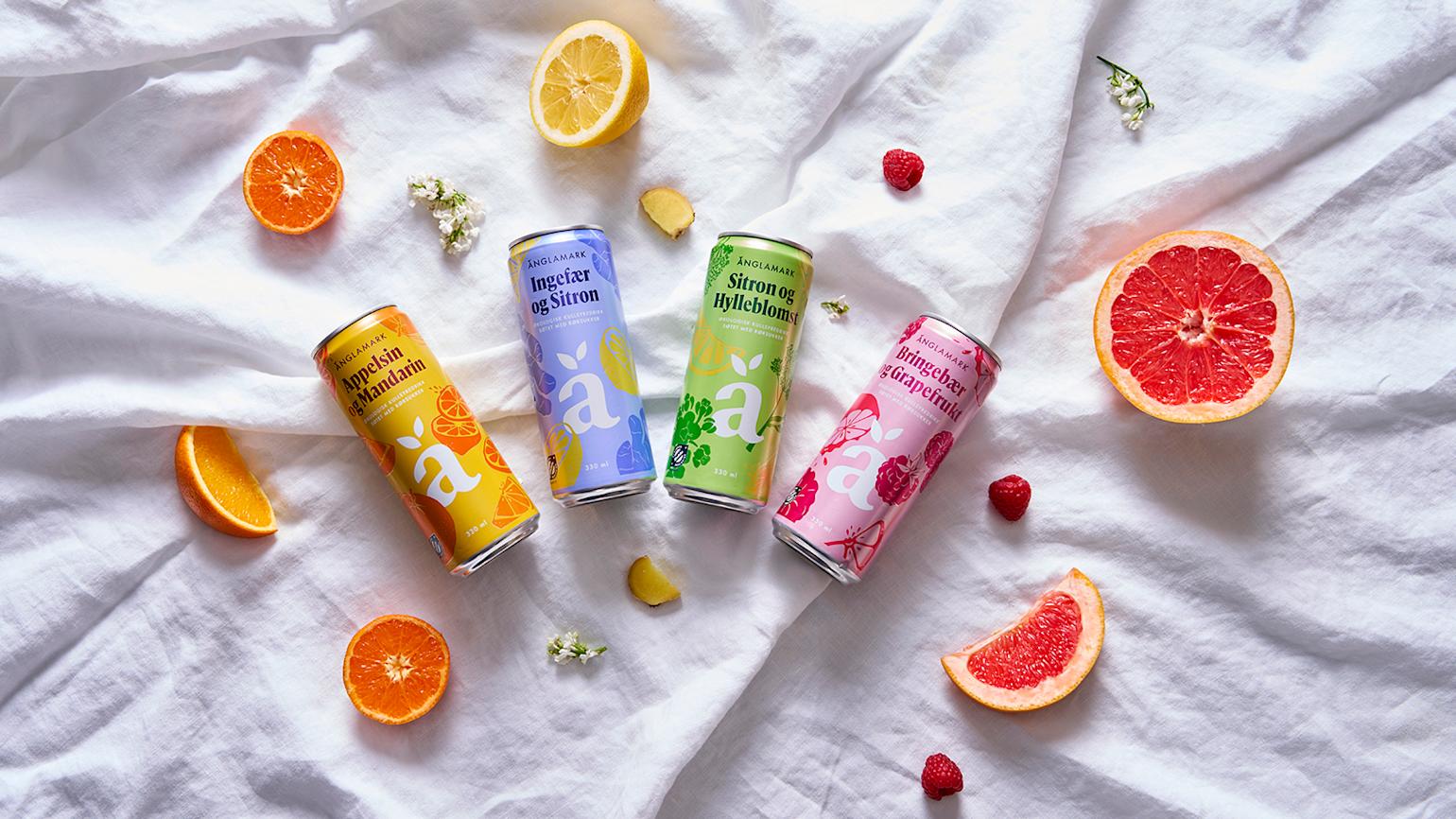 Änglamark drikke i alle fire varianter liggende på hvit linduk med frisk frukt, bær og blomster rundt.