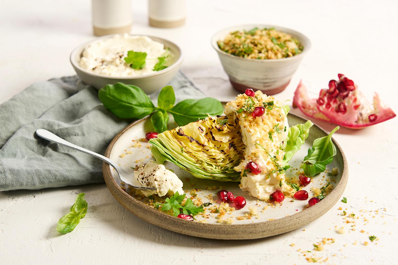 Grillet nykål med fetakrem, urtecrunch og granateple servert på lys keramikktallerken med friske basilikumblader og en skje med fetakrem på kanten. Skåler med fetakrem og urtecrunch i bakgrunnen.