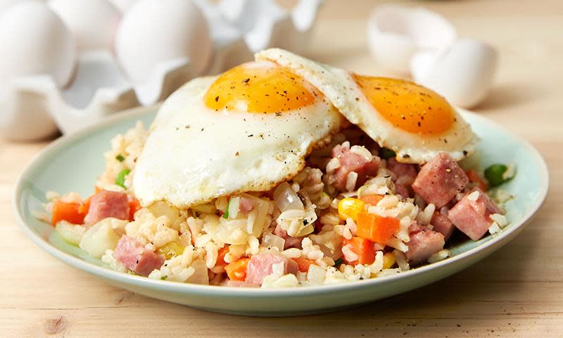 Stekt ris med skinke og speilegg, amerikansk grønnsaksblanding og løk. Servert på lys grønn tallerken, på lyst trebord. Hele egg i bakgrunnen.