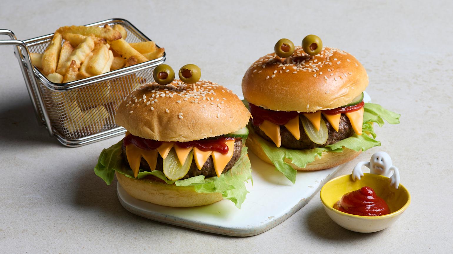 Halloween hamburgere laget som monstere med ost, salat, pickles og oliven. Liten skål med ketchup og et spøkelse på siden.