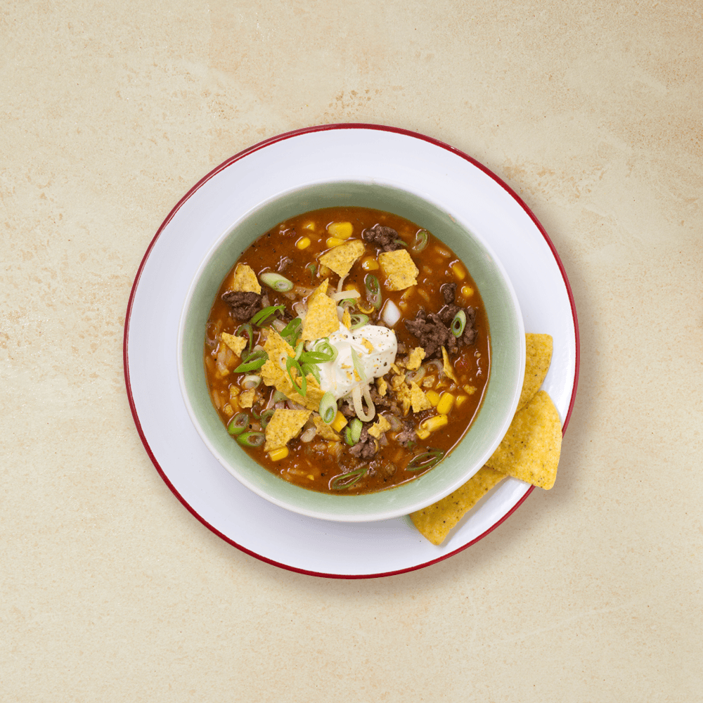 Meksikansk tacosuppe toppet med vårløk, nacho chips, revet ost og rømme i en grønn skål på en hvit tallerken. Servert med nacho chips.