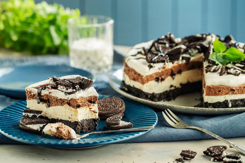 Oreokake i tre lag med kjeksbunn, ostekrem og sjokolademousse. Pyntet med pisket krem, knuste Oreokjeks, sjokoladesaus og noen blader sitronmelisse. Ett stykke servert på blå asjett ved siden av kaken.