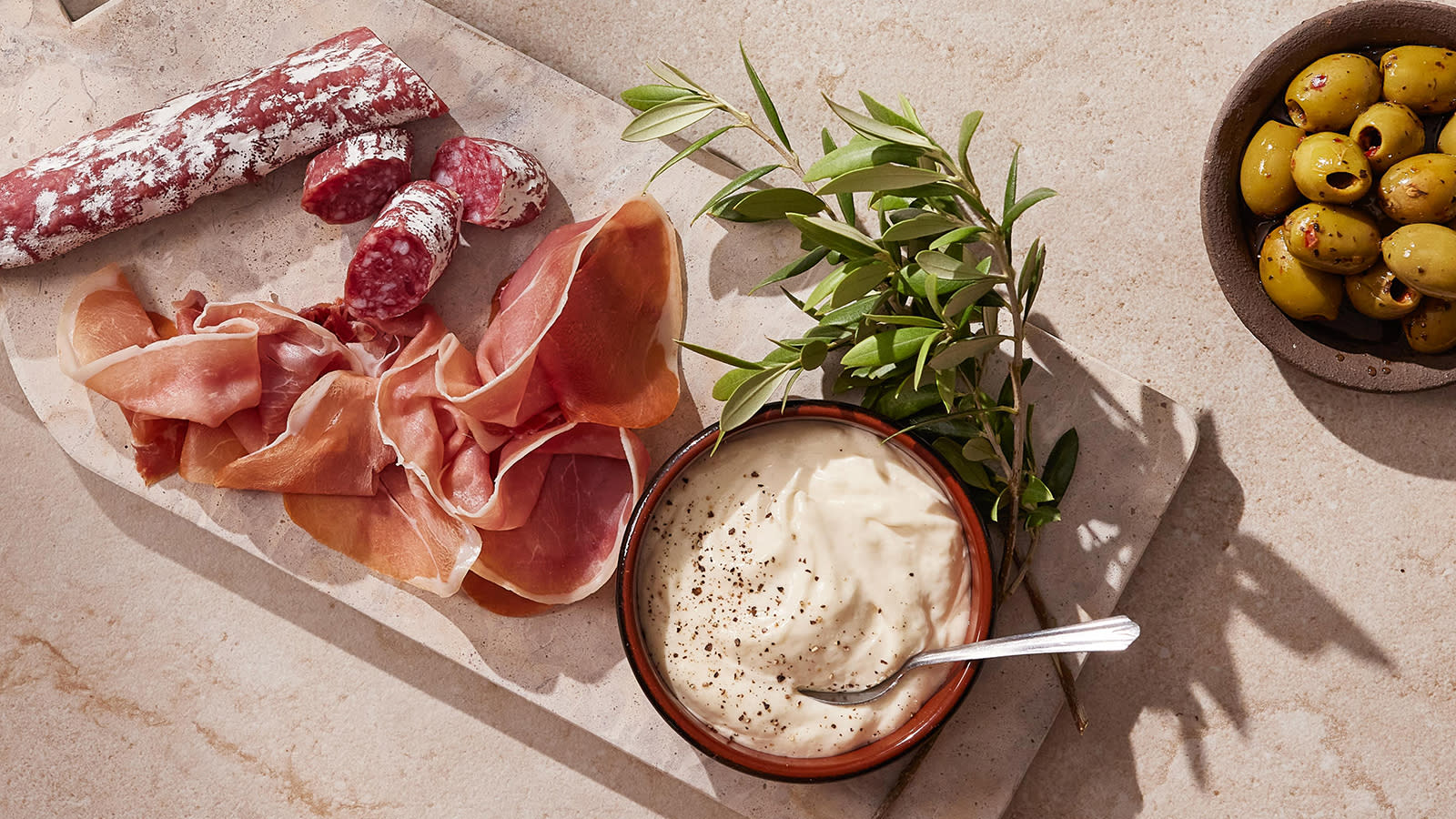 Rask hjemmelaget aioli servert på marmorfat med spekeskinke og spekepølse. Liten skål med grønne oliven på siden.