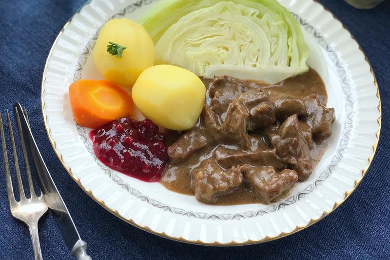 En pen hvit middagstallerken med sosekjøtt, servert med kokt potet, gulrot, kål og tyttebærsyltetøy.