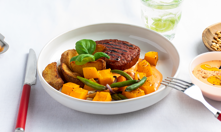 Vegansk biff med ovnsbakte poteter og grønnsaker servert i en hvit skål med bestikk med rødt håndtak, og et glass vann med en limebåt i bakgrunnen