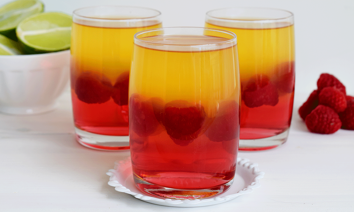 Sukkerfri gelé i to lag med bringebær mellom, servert i dessertglass på hvitt bord. Ferske bringebær, og liten skål med limebåter i bakgrunnen.