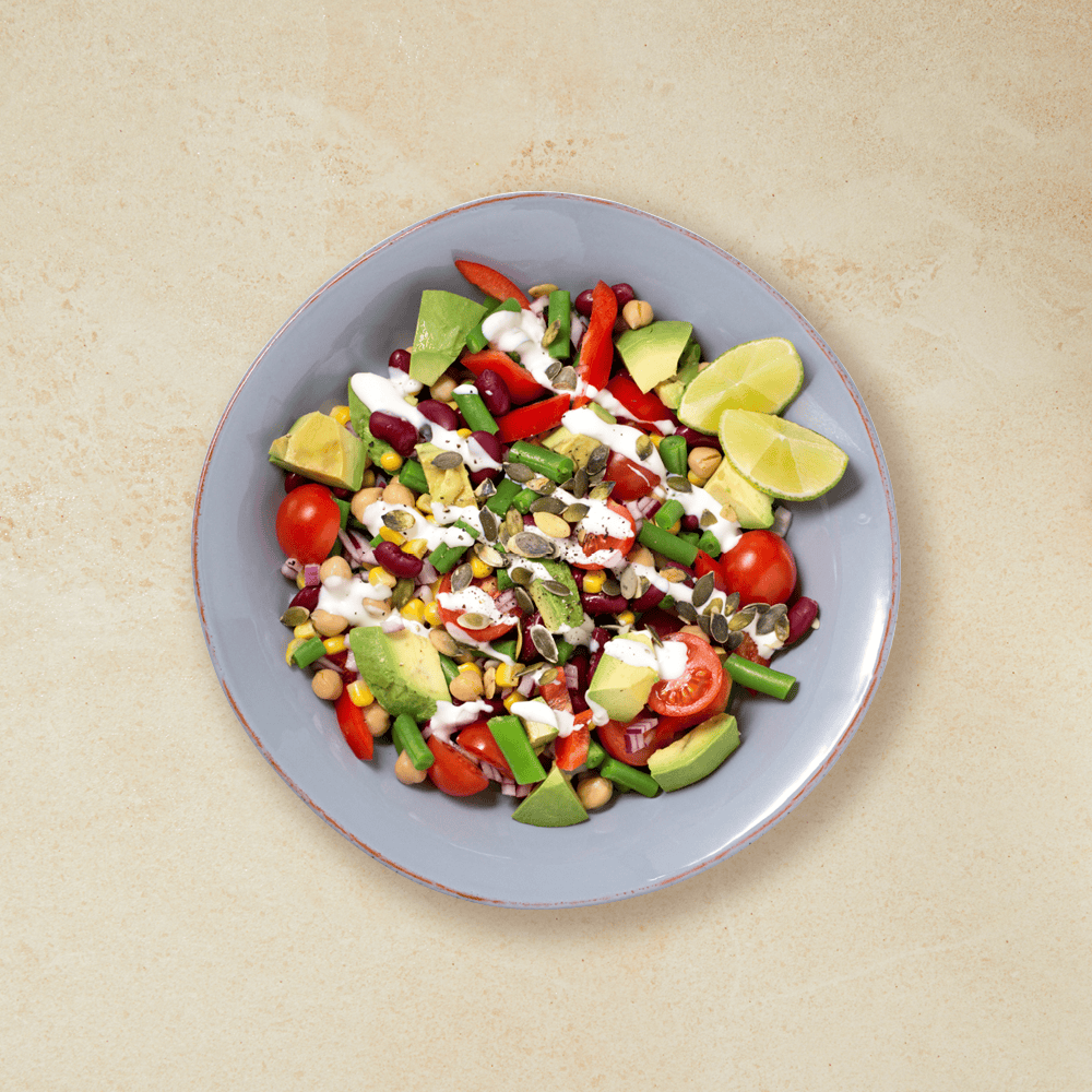 Vegetarsalat med kikerter, bønner, avokado, tomater, paprika og gresskarkjerner. Limebåter på siden.