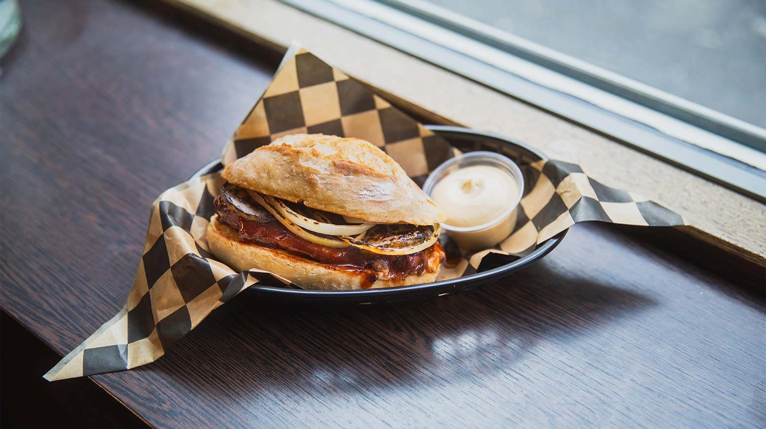 The Render Rib med Grill Perfekt baby back ribs servert i ciabatta i burgerkurv i en vinduskarm. Liten skål med dip ved siden av i kurven.