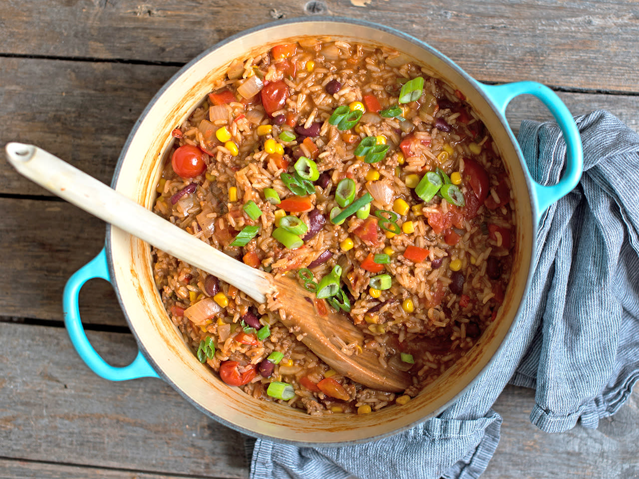 Gryterett med meksikansk vri, med tomat, bønner, mais, kjøttdeig og vårløk, laget i turkis kjele. Rustikk trebord og blått kjøkkenhåndkle ved siden av.