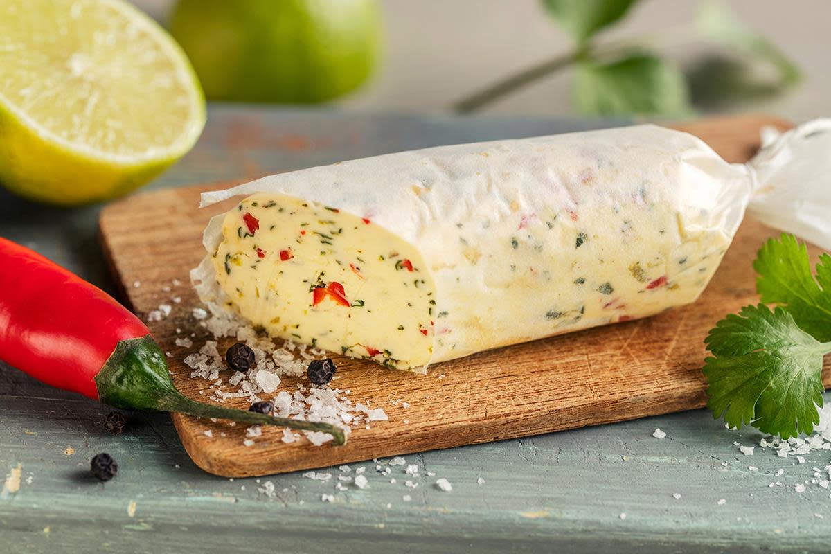 Kryddersmør med chili og lime, rullet til en pølse og pakket i hvitt papir, servert på liten trefjøl med ferske urter rundt.