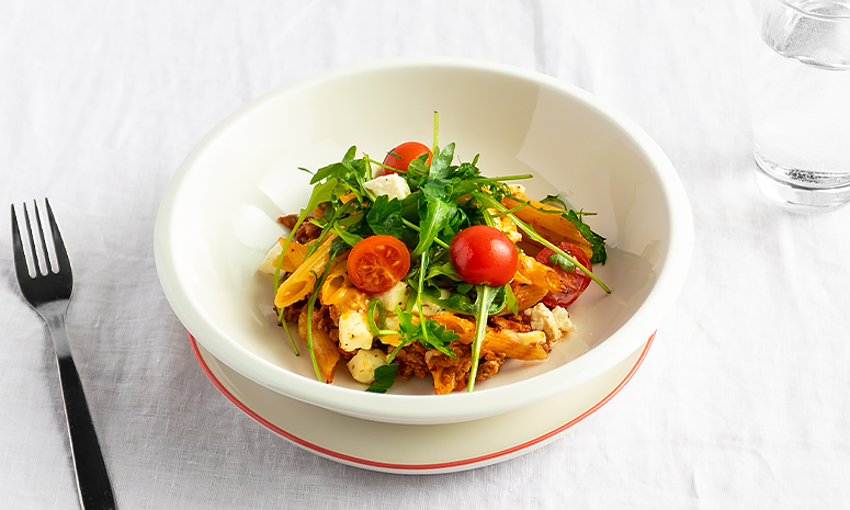 Kjøttdeig og pasta i form servert i en hvit skål som står på en liten hvit tallerken med en gaffel liggende på venstre side, alt ligger på en hvit duk