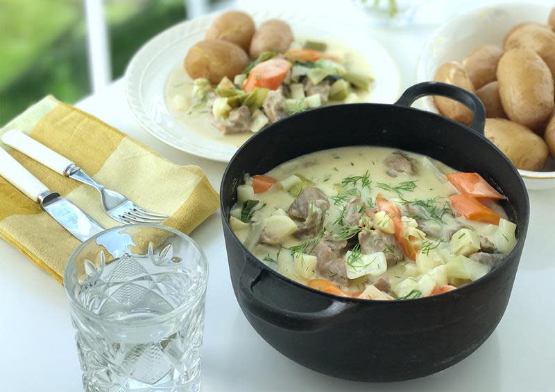 Kremet lammefrikassé med gulrøtter og purre, servert i en sort støpejernsgryte. Kokte poteter på siden. Lys bakgrunn.