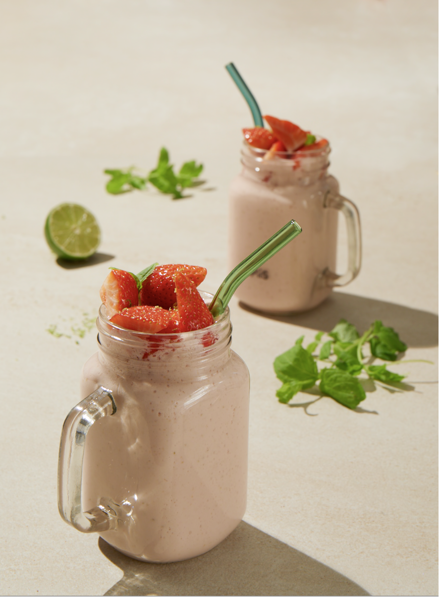 Lys rosa jordbærmilkshake i smoothieglass med sugerør, på lys bakgrunn. Ferske jordbær på toppen, og ferske urter, og lime rundt glassene.