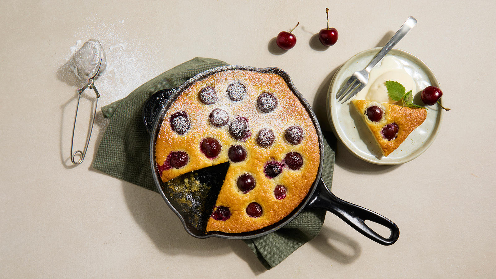 Morellcobbler - enkel dessertpai med moreller, servert i rund støpejernspanne på grønt kjøkkenhåndkle. Liten asjett med et stykke kobler og vaniljeis på siden.