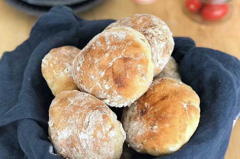 Enkle eltefrie rundstykker med et dryss mel, servert i kurv med mørkeblå linserviett på trebord.