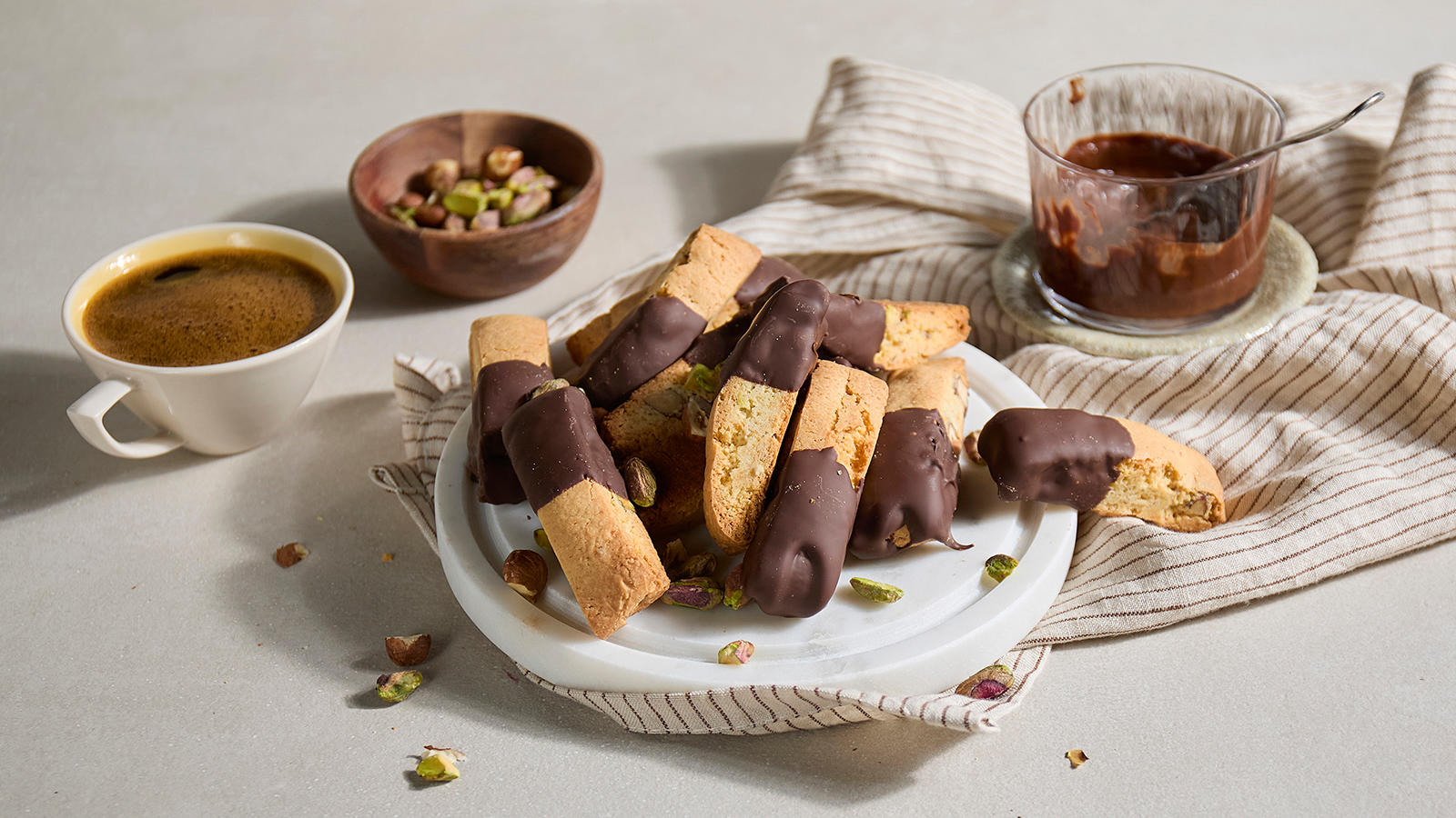 Biscotti med sjokolade og pistasj servert på rund marmorasjett med en kopp kaffe ved siden av. Liten skål med smeltet sjokolade på siden, og skål med pistasjenøtter i bakgrunnen.