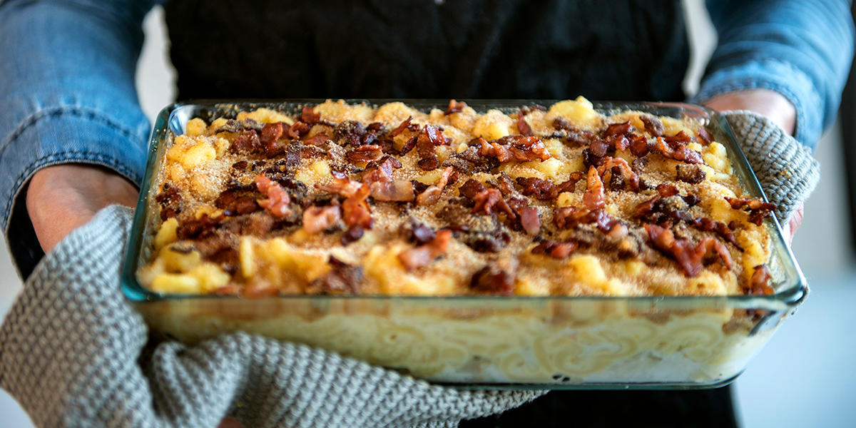 Mac and cheese med torsk og bacon servert i gjennomsiktig ildfast form av glass holdt frem av to armer med strikket kjøkkenhåndkle.