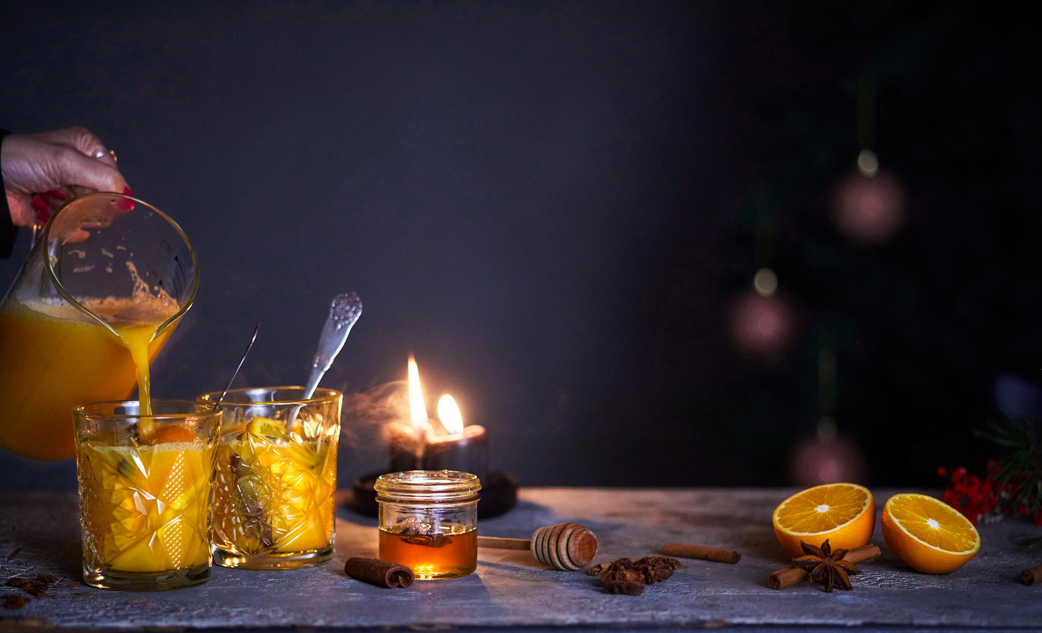 Appelsingløgg med julekrydder servert i to glass på skiferhelle i mørkt rom med tente lys, hele tørkede krydder og delt fersk appelsin. Appelsingløgg helles i glassene av en mugge i glass.