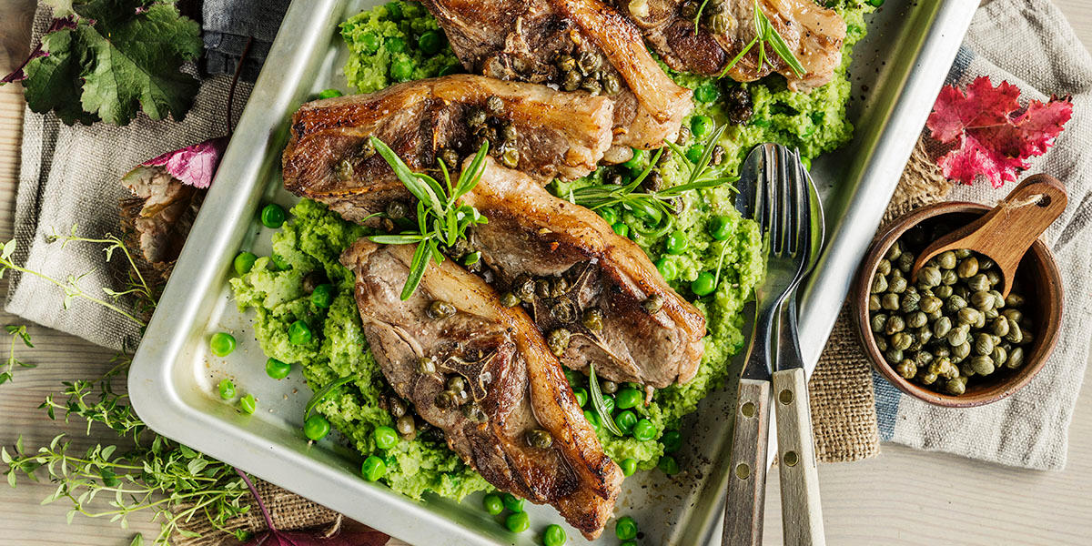 Sitronstekte lammekoteletter servert på en seng grønn potetmos med erter. Ferske urter og kapers er strødd over. Fatet står på et lyst trebord med linservietter, og en liten treskål med kapers ved siden av.