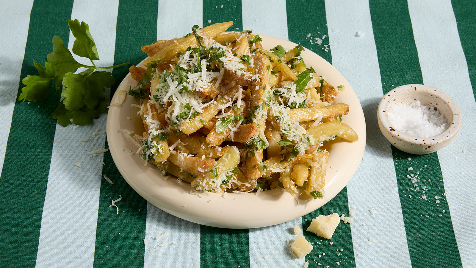 Parmesan fries servert på beige tallerken på grønnstripet duk. Friske urter og en skål med flaksalt ved siden av.