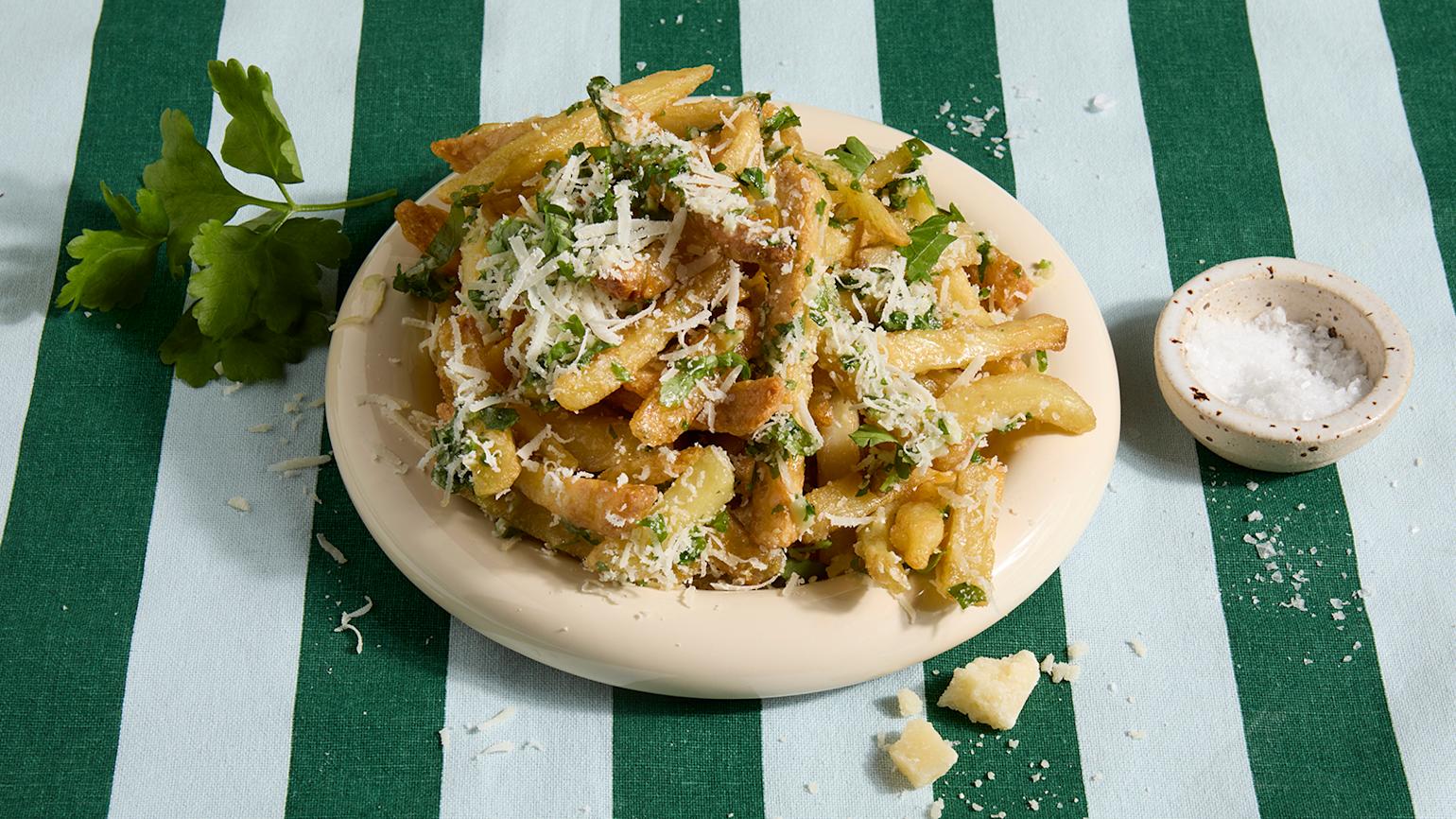 Parmesan fries servert på beige tallerken på grønnstripet duk. Friske urter og en skål med flaksalt ved siden av.