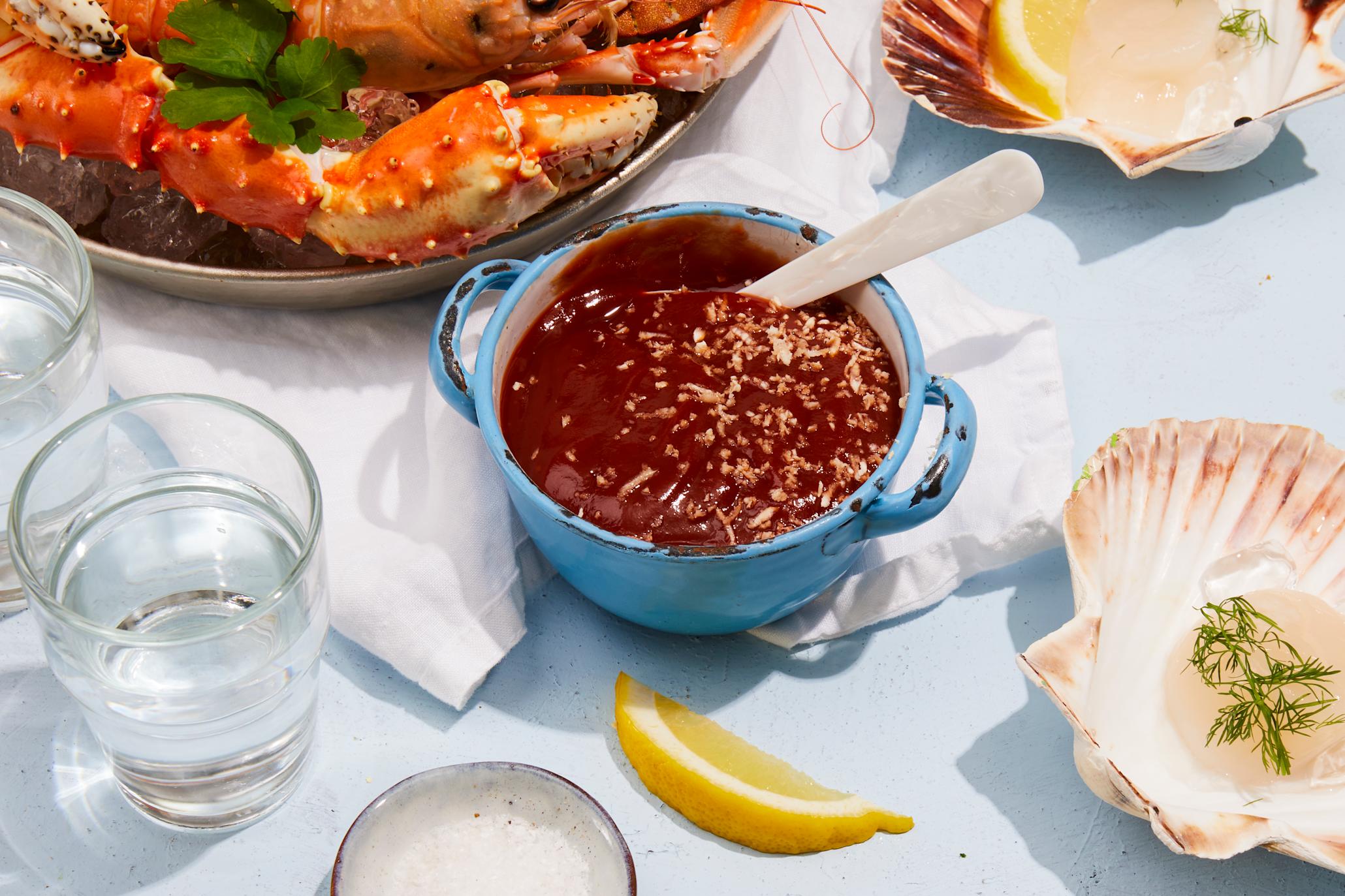Cocktailsaus til skalldyr servert i blå skål med små håndtak, på dekket skalldyrbord med sitronskiver, vannglass, sjøkreps, krabbe og kamskjell.