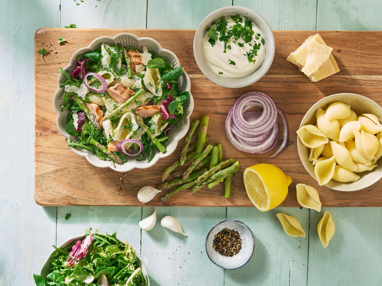 Pastasalat med grillet laks, asparges, rødløk, dressing og parmesan servert i lys skål på trefjøl med friske grønnsaker rundt.