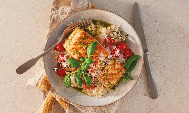 Laks med pesto servert over hjemmelaget sitronrisotto og bakte tomater i lys rustikk keramikkskål med beige og gul linserviett under. Laksen er pyntet med ferske basilikumblader og revet parmesan.