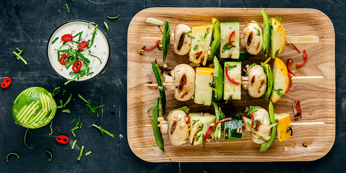 Bildet er tatt ovenfra og viser grillspyd med squash, sopp, grønn paprika, sukkererter, urter og chili. Ved siden av står en skål med rømmedipp med revet limeskall og chili.