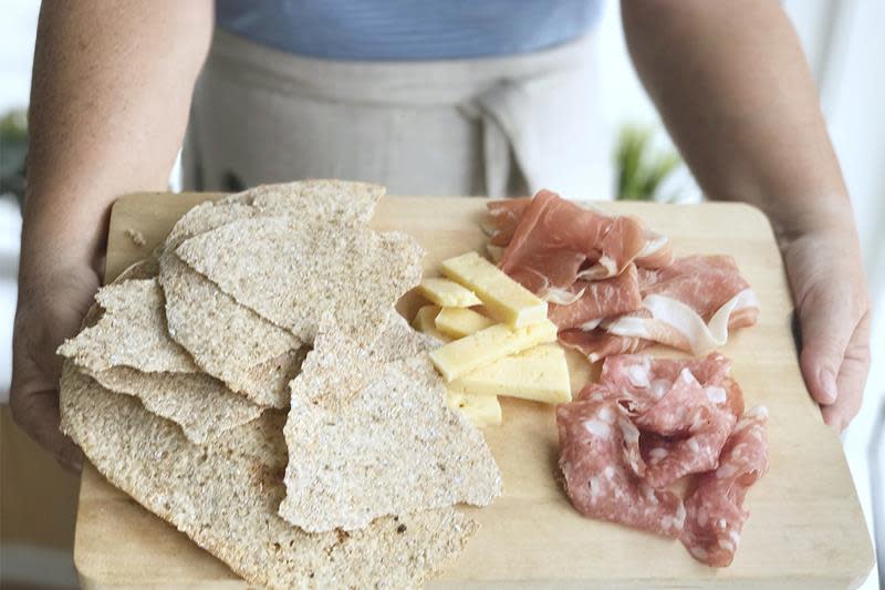 Hjemmelaget flatbrød servert på lys trefjøl med spekemat og ost, holdes frem av to hender.