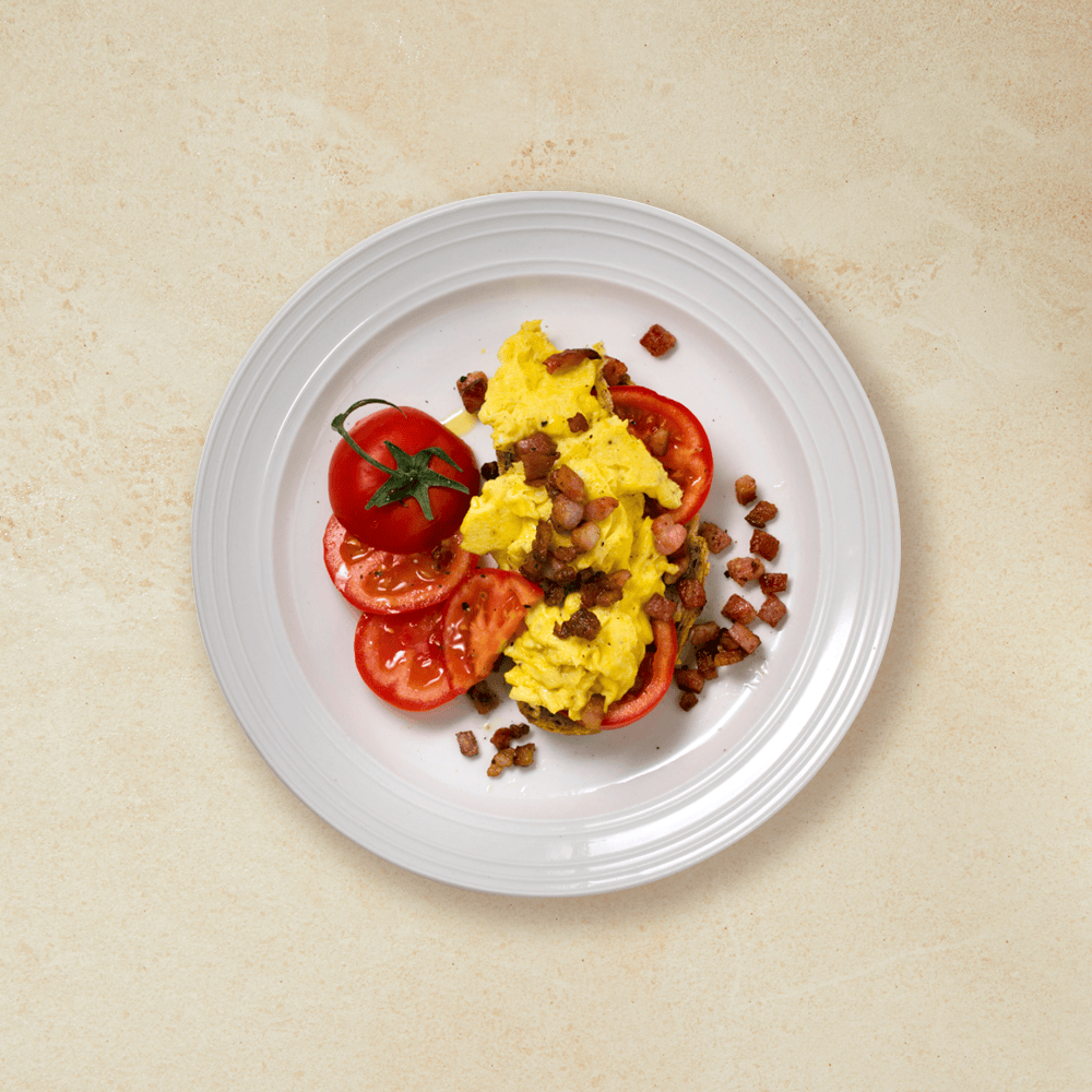 Eggerøre på en skive brød med tomatskiver og smårettbacon servert på hvit tallerken, på beige bakgrunn.