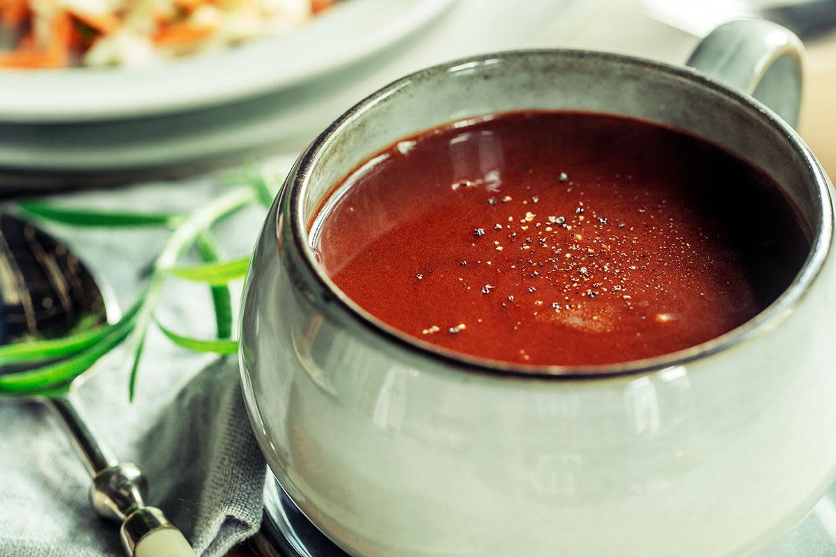 Mørk rødbrun rødvinssaus til lam, toppet med nykvernet pepper. Servert i gråmelert sausemugge, på bord, med grå linserviett og ferske urter ved siden av.